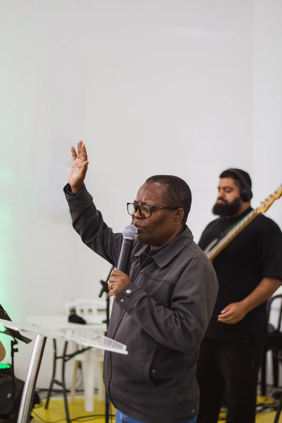 Homem de mão levantada conduzindo a oração ao microfone, com o baixista da banda ao fundo.