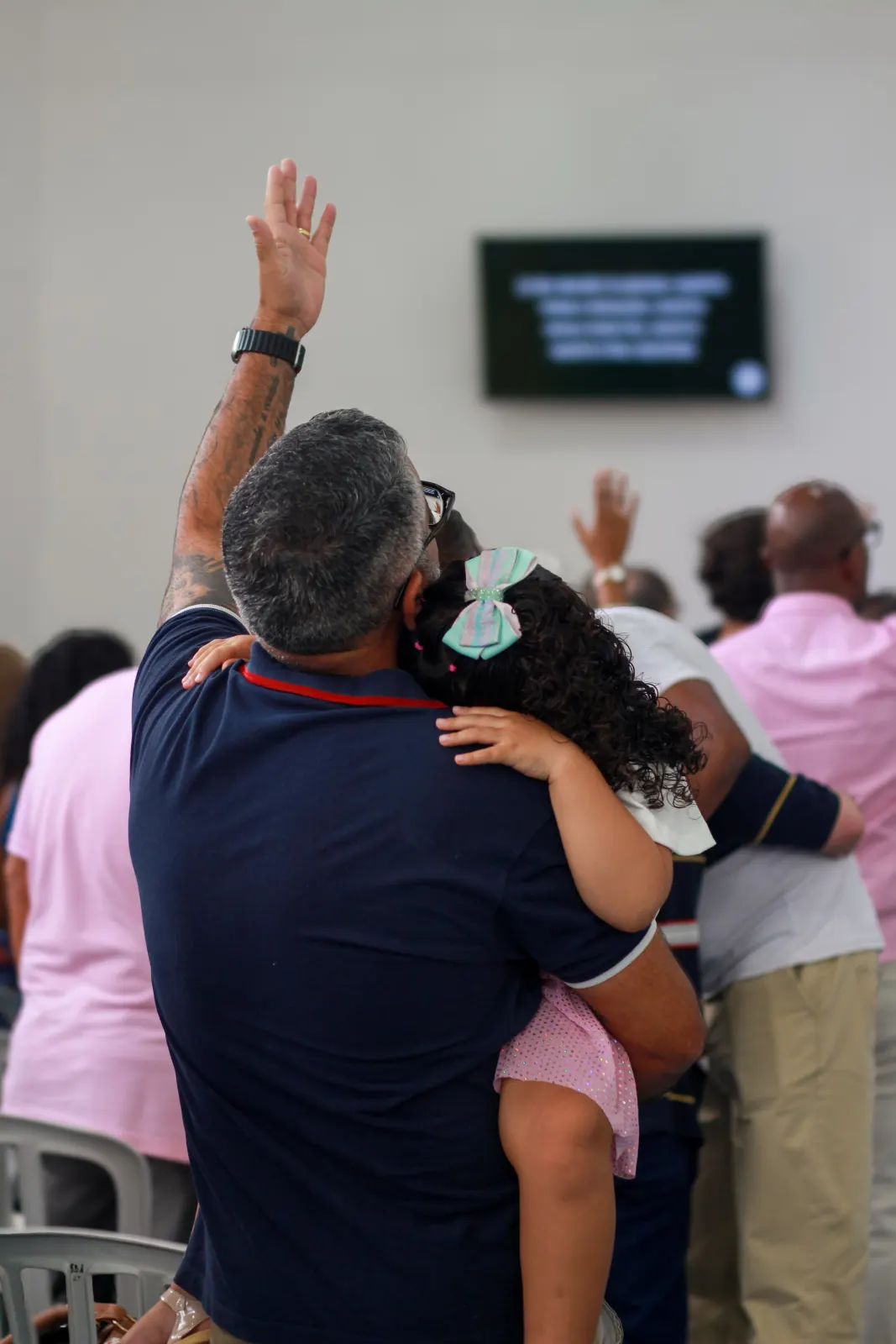 Pai com a filha no colo, de mão erguida durante um momento de louvor, cercado por outros membros também com as mãos levantadas.